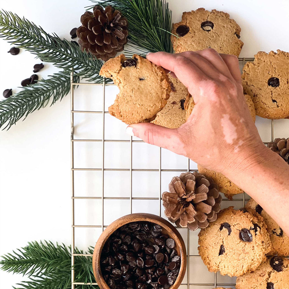 
                  
                    A photo of a hand placing a chocolate chip cookie with a bite-sized chunk missing onto a wire cooling rack, that already had a number of cookies already on it, along with some decorative pine cones, some pine branches, and a bowl of dark chocolate chips.
                  
                